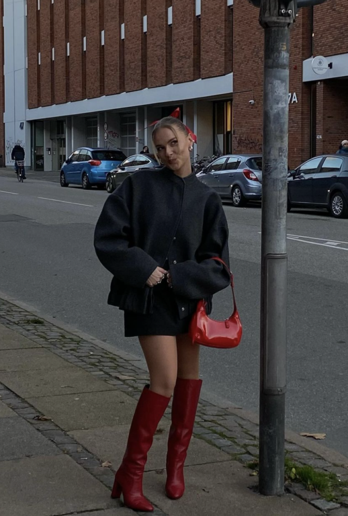 Woman wearing a black dress with a cozy oversized jacket, red knee-high boots, and a matching red handbag, standing on a city street.